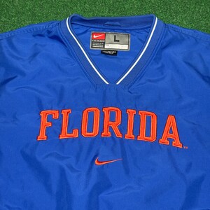 以下が含まれることがあります： 胸にオレンジ色のブロック体で「FLORIDA」の文字が入ったロイヤルブルーのプルオーバージャケット。Vネックの襟には白いトリム、小さなオレンジ色のナイキのスウッシュロゴが付いています。