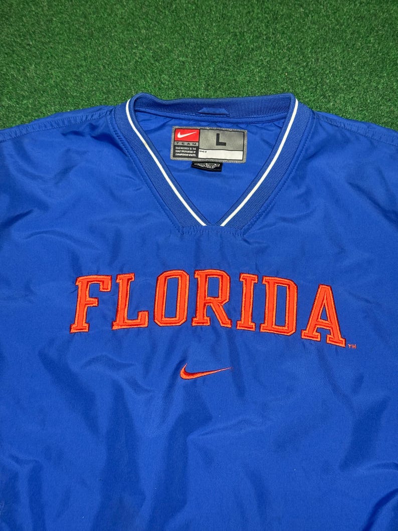 以下が含まれることがあります： 胸にオレンジ色のブロック体で「FLORIDA」の文字が入ったロイヤルブルーのプルオーバージャケット。Vネックの襟には白いトリム、小さなオレンジ色のナイキのスウッシュロゴが付いています。