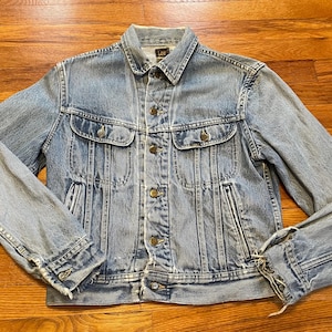 Könnte beinhalten: Eine hellblaue Jeansjacke mit Knopfleiste, zwei Brusttaschen und zwei Fronttaschen. Die Jacke hat einen ausgewaschenen Look und ist leicht abgenutzt.