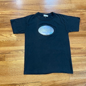 Puede incluir: Una camiseta negra con un logotipo de Batman plateado y reflectante en la parte delantera.