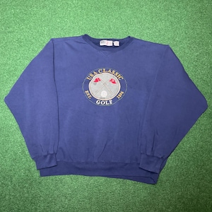 May include: Navy blue sweatshirt with a crew neck. The front features a circular embroidered design with the text "USA Classic Golf Est. 1996". The design includes crossed golf clubs, a golf ball, and two red flags. The sweatshirt has ribbed cuffs and hem.