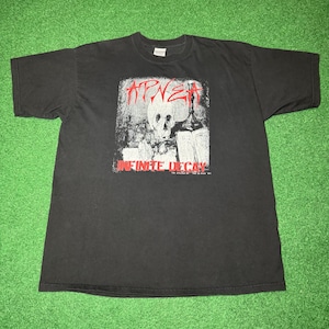 May include: Black t-shirt featuring a graphic design with a skull, bottle, and text. The text includes "APNEA" in red, and "INFINITE DECAY" in red. The shirt is laid flat on a green surface.