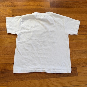 Może przedstawiać: Gładki biały t-shirt z kr&oacute;tkim rękawem, widok z tyłu. Koszulka wykonana jest z miękkiego materiału, prawdopodobnie bawełny. Koszulka leży na drewnianej powierzchni.