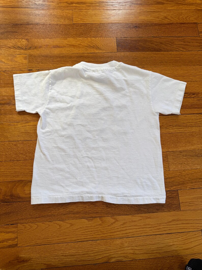 Może przedstawiać: Gładki biały t-shirt z kr&oacute;tkim rękawem, widok z tyłu. Koszulka wykonana jest z miękkiego materiału, prawdopodobnie bawełny. Koszulka leży na drewnianej powierzchni.