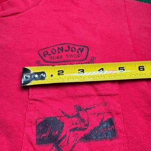 May include: Red t-shirt with a black graphic of a surfer riding a wave. The shirt has a pocket on the front with a black logo that reads "Ronjon Surf Shop".