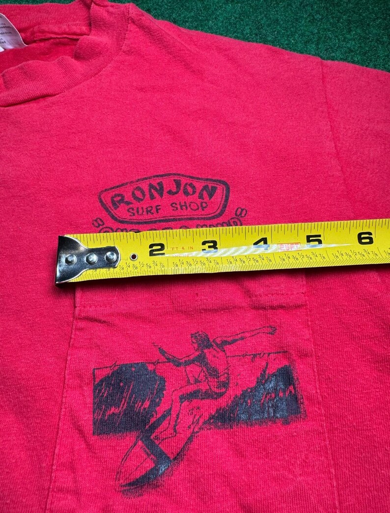 May include: Red t-shirt with a black graphic of a surfer riding a wave. The shirt has a pocket on the front with a black logo that reads "Ronjon Surf Shop".