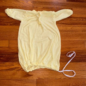May include: A pale yellow long-sleeved gown featuring a round neckline with a single button closure at the back. The bottom hem is gathered with an elastic band and includes a long white drawstring tie extending from the right side of the hem.