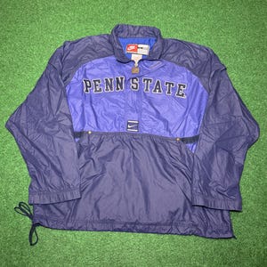 Puede incluir: Chaqueta tipo jersey Nike Penn State azul marino y morado. La chaqueta presenta las palabras "PENN STATE" en letras blancas en el pecho. Tiene cierre de cremallera de un cuarto, un pequeño logotipo de Nike y un cordón en el dobladillo inferior.