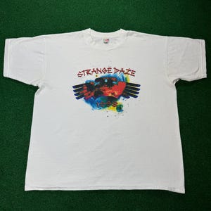 May include: White t-shirt with a colorful graphic print of a hand with wings and the text "Strange Daze 1988".