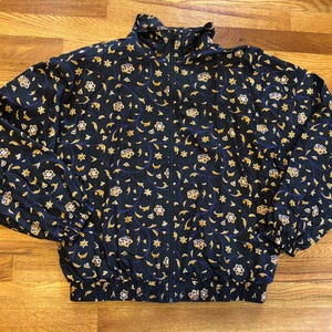 Puede incluir: Una chaqueta de satén negra con un estampado floral dorado y morado. La chaqueta tiene una cremallera y un cuello de pie.