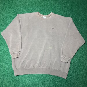 May include: A faded brown crewneck sweatshirt with the Nike logo embroidered in black on the chest. The sweatshirt has a small Nike tag on the neckline and ribbed cuffs and hem. The garment is made of a soft, worn material.
