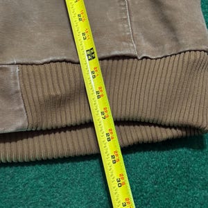 Puede incluir: Una sudadera con capucha marr&oacute;n con dobladillo inferior acanalado. Una cinta m&eacute;trica amarilla se coloca sobre la prenda, mostrando medidas de hasta 81 cm.