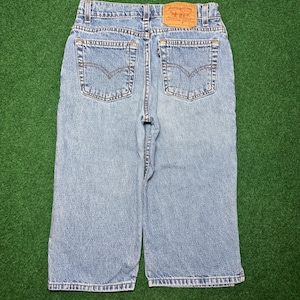 May include: Light blue denim jeans featuring a high-waisted cut, two back pockets with signature arcuate stitching, a tan leather patch on the waistband, and a small black tab on the right pocket. The jeans have a straight leg and classic metal rivets.