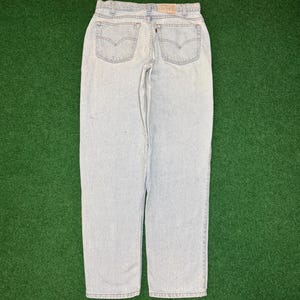 May include: Light wash denim jeans with classic five-pocket styling. The jeans feature the iconic Levi's back pocket design and a leather patch at the waistband. The jeans are displayed on a green surface.