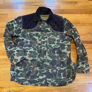 May include: Camouflage pattern jacket with a black quilted shoulder panel and collar. The jacket has multiple pockets and button closures. The camouflage design features shades of green, brown, and tan.