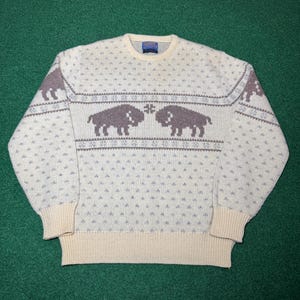 May include: A cream-colored crew neck sweater featuring a repeating pattern of brown bison silhouettes across the chest and shoulders. The knit garment includes light blue geometric accents, ribbed cuffs, and a ribbed hem, with a Pendleton brand label.