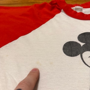 Pu&ograve; includere: T-shirt bianco e rosso in stile raglan con grafica di Topolino nero. La maglietta ha colletto e maniche rosse e corpo bianco. L'immagine &egrave; un primo piano della parte anteriore della maglietta, con un dito che indica il tessuto.