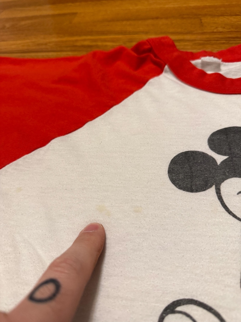 Pu&ograve; includere: T-shirt bianco e rosso in stile raglan con grafica di Topolino nero. La maglietta ha colletto e maniche rosse e corpo bianco. L'immagine &egrave; un primo piano della parte anteriore della maglietta, con un dito che indica il tessuto.