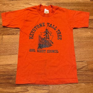 May include: An orange t-shirt with a crew neck and short sleeves. The front of the shirt features a graphic with the text "Keystone Tall Tree" around a tree illustration, and "Girl Scout Council" below.
