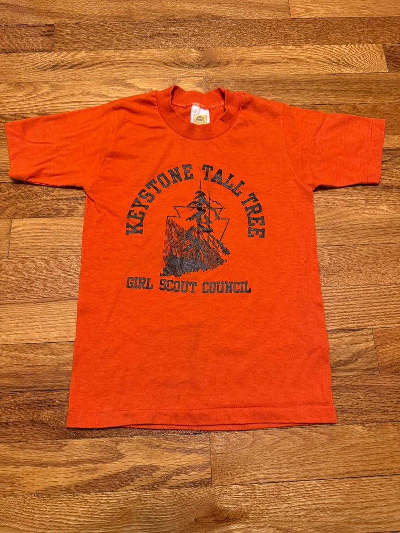 May include: An orange t-shirt with a crew neck and short sleeves. The front of the shirt features a graphic with the text "Keystone Tall Tree" around a tree illustration, and "Girl Scout Council" below.