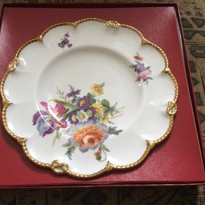 Puede incluir: Un plato de porcelana blanca con un borde dorado y un diseño floral. El plato está en una caja roja.