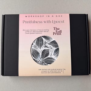 Può includere: Una scatola nera con la scritta "Workshop in a Box" e il titolo "Printfulness with Linocut". La scatola presenta un motivo floreale in bianco e nero. Il testo descrive un workshop per imparare la linoleografia rilassandosi con una meditazione audio.