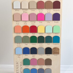 May include: A rectangular wooden display board titled By His Saving Grace Paint Options, featuring rows of arched paint swatches in various colors including white, pink, blue, green, and metallic tones, each labeled with its corresponding color name.