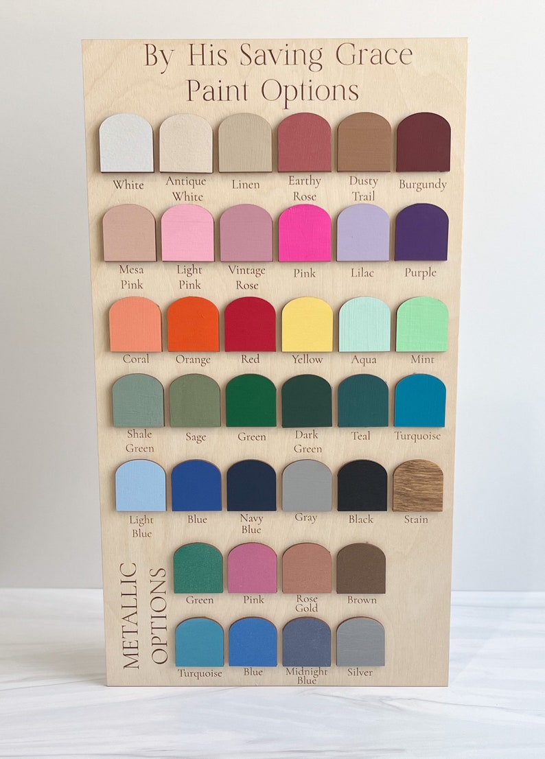 May include: A rectangular wooden display board titled By His Saving Grace Paint Options, featuring rows of arched paint swatches in various colors including white, pink, blue, green, and metallic tones, each labeled with its corresponding color name.