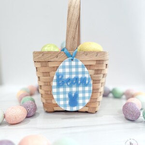 May include: A woven basket with a wooden handle, filled with yellow and green eggs. A blue and white gingham Easter egg tag with the name "Beau" and a bunny is attached to the basket with a blue ribbon. Pastel-colored Easter eggs surround the basket.