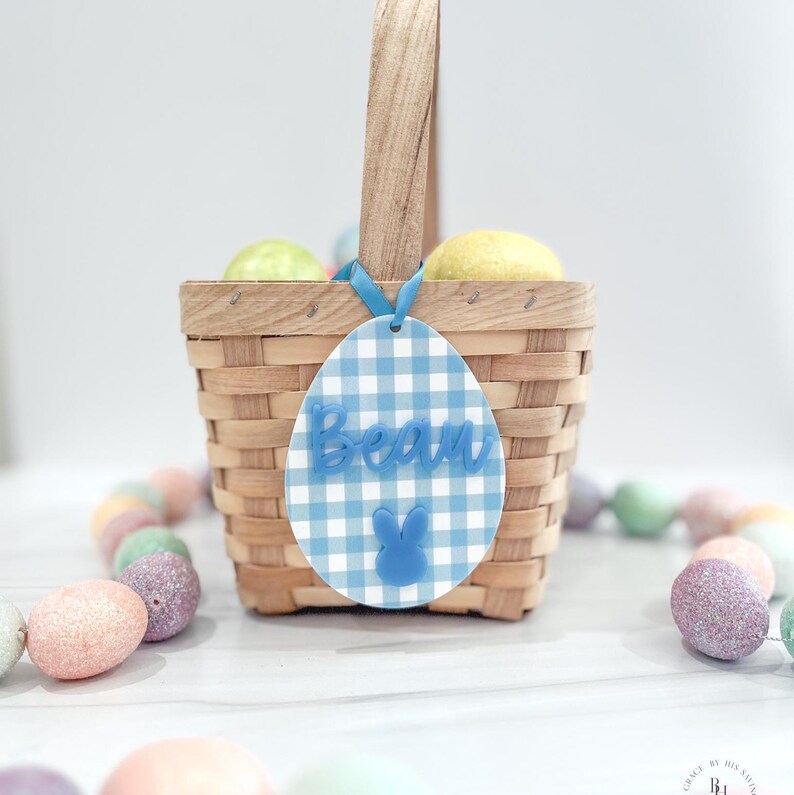 May include: A woven basket with a wooden handle, filled with yellow and green eggs. A blue and white gingham Easter egg tag with the name "Beau" and a bunny is attached to the basket with a blue ribbon. Pastel-colored Easter eggs surround the basket.