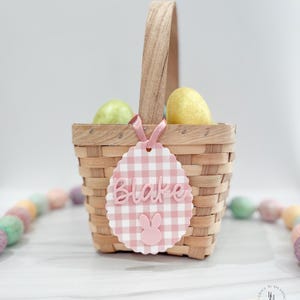 May include: A woven basket with a wooden handle, holding two decorative eggs. A pink and white gingham tag with the name "Blake" and a bunny is attached to the basket with a pink ribbon. Pastel-colored Easter eggs are in the background.