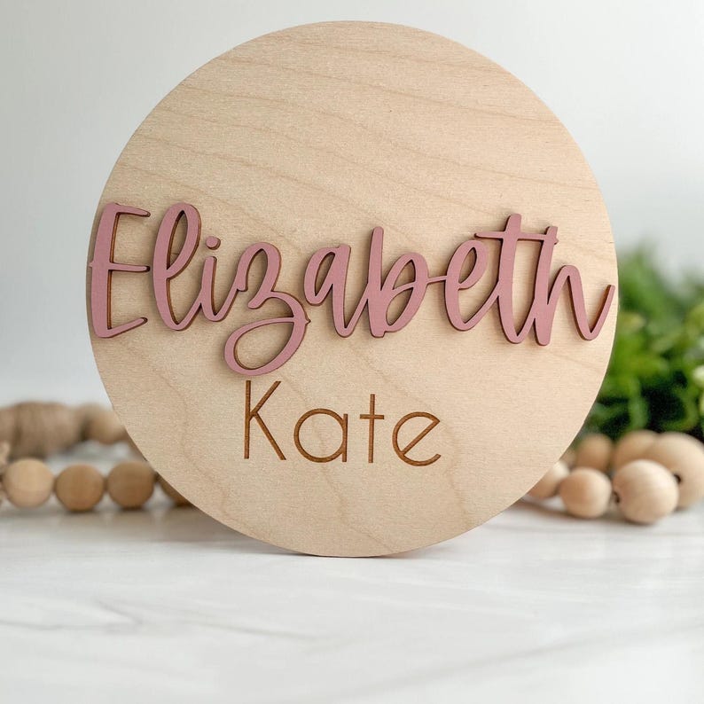 May include: A circular light-colored wooden plaque featuring the name Elizabeth in raised, dusty rose-colored script lettering and the name Kate engraved in a simple, dark brown sans-serif font centered below the script text.