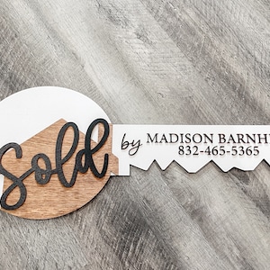 May include: A white and brown wooden key-shaped sign with the word "Sold" in black lettering on the key head and the text "by MADISON BARNHILL 832-465-5365" on the key blade.