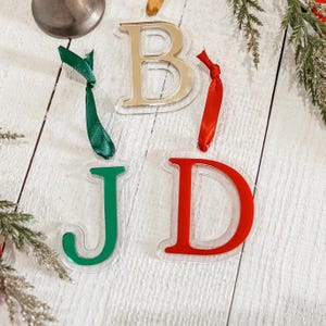 May include: Acrylic Christmas ornaments in the shape of the letters B, J, and D. The letter B is gold, J is green, and D is red. Each letter has a ribbon for hanging. The ornaments are on a white wooden surface with greenery.