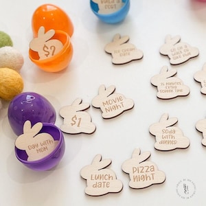 May include: An overhead shot shows colorful Easter eggs and wooden bunny-shaped tokens. The eggs are orange, purple, and blue, some open with tokens inside. The tokens have phrases like "Movie Night" and "Lunch Date."