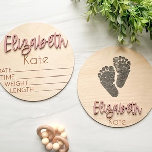 May include: Two circular light-colored wooden plaques featuring the name Elizabeth Kate in raised dusty pink lettering. One plaque includes engraved lines for date, time, weight, and length. The other plaque features a dark engraved footprint design.
