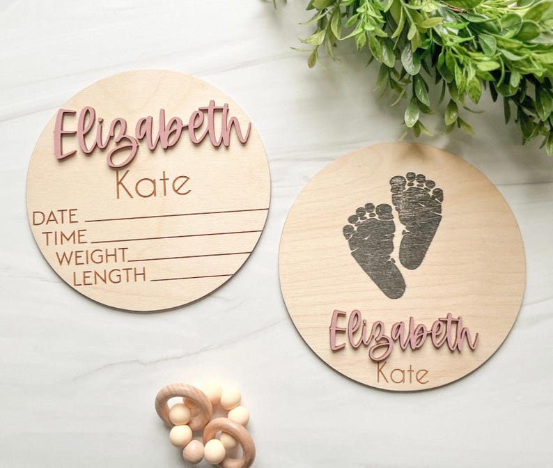 May include: Two circular light-colored wooden plaques featuring the name Elizabeth Kate in raised dusty pink lettering. One plaque includes engraved lines for date, time, weight, and length. The other plaque features a dark engraved footprint design.