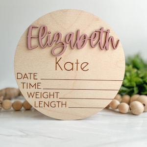 May include: A circular light wood plaque featuring the name Elizabeth Kate in raised dusty pink script lettering. Below the name, the words DATE, TIME, WEIGHT, and LENGTH are engraved in a simple font, each followed by a horizontal line for writing.