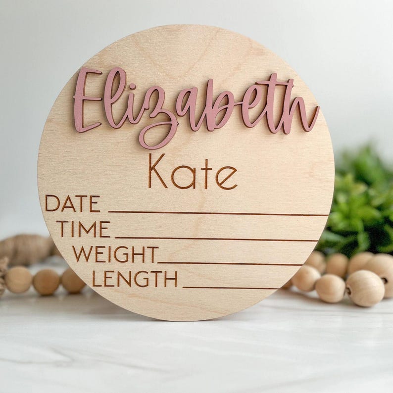 May include: A circular light wood plaque featuring the name Elizabeth Kate in raised dusty pink script lettering. Below the name, the words DATE, TIME, WEIGHT, and LENGTH are engraved in a simple font, each followed by a horizontal line for writing.