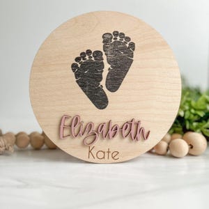 May include: A circular light-colored wooden plaque featuring a dark brown printed graphic of two baby footprints. The name Elizabeth is displayed in raised, dusty pink script lettering, with the name Kate printed below in brown text.