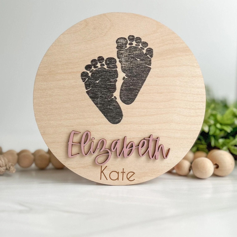 May include: A circular light-colored wooden plaque featuring a dark brown printed graphic of two baby footprints. The name Elizabeth is displayed in raised, dusty pink script lettering, with the name Kate printed below in brown text.