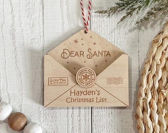 Puede incluir: Un adorno de madera clara con forma de sobre que incluye el texto Dear Santa, Sleigh Mail Special Delivery y Hayden's Christmas List. Un sello circular de North Pole Post está centrado en la solapa, con un lazo de cuerda roja y blanca.