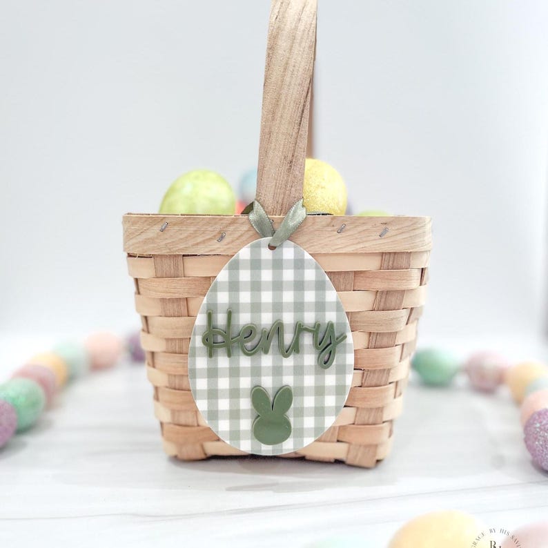 May include: A woven basket with a wooden handle, filled with colorful Easter eggs. An egg-shaped tag with the name "Henry" in green lettering and a green bunny graphic hangs from the handle. The tag has a gray and white gingham pattern.