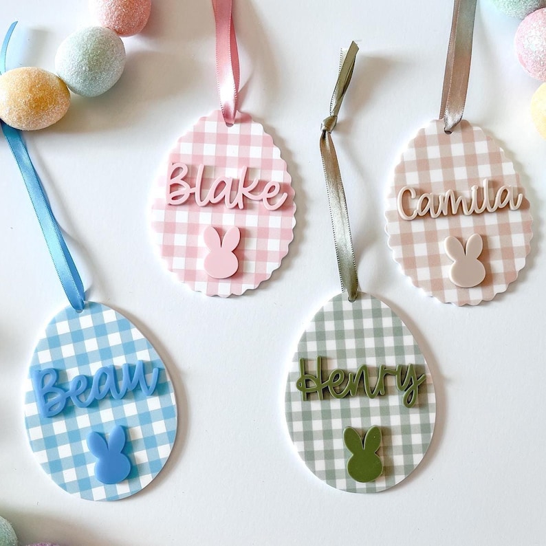 May include: Easter egg-shaped name tags with gingham patterns in pink, blue, and green. Each tag features a name and a bunny cutout. Ribbons in coordinating colors are attached for hanging. Decorative Easter eggs are in the background.