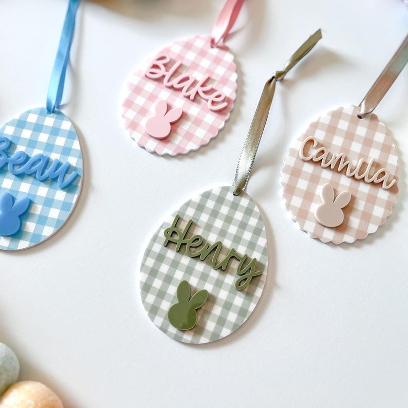 May include: Easter egg-shaped name tags with gingham patterns in blue, pink, and green. Each tag features a name and a bunny silhouette. Ribbons in coordinating colors are attached for hanging. A festive and personalized touch for Easter.