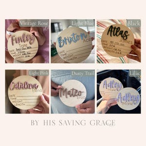 May include: A collection of six circular wooden birth announcement signs featuring raised 3D lettering in various colors including vintage rose, light blue, black, light pink, dusty trail, and lilac, with printed lines for date, time, weight, and length.