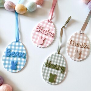 May include: Easter egg-shaped name tags with gingham patterns in blue, pink, and green. Each tag features a name and a bunny silhouette. Ribbons in coordinating colors are attached for hanging. A festive Easter decoration.