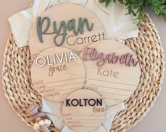 May include: Four circular wooden birth announcement signs with raised lettering in sage green, white, dusty rose, and navy blue. Each sign features the names Ryan Garrett, Olivia Grace, Elizabeth Kate, and Kolton Lane with blank lines for date, time, weight, and length.