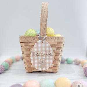 May include: A woven basket with a wooden handle, filled with colorful Easter eggs. A decorative tag with the name "Camila" and a bunny design hangs from the handle. The tag has a scalloped edge and a gingham pattern.