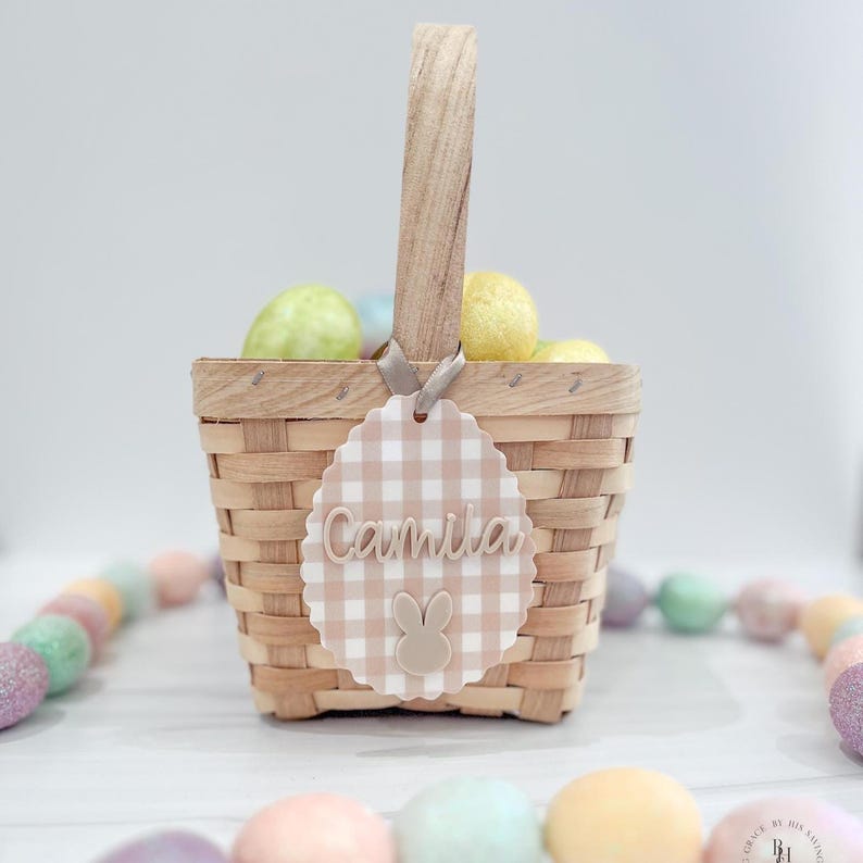 May include: A woven basket with a wooden handle, filled with colorful Easter eggs. A decorative tag with the name "Camila" and a bunny design hangs from the handle. The tag has a scalloped edge and a gingham pattern.